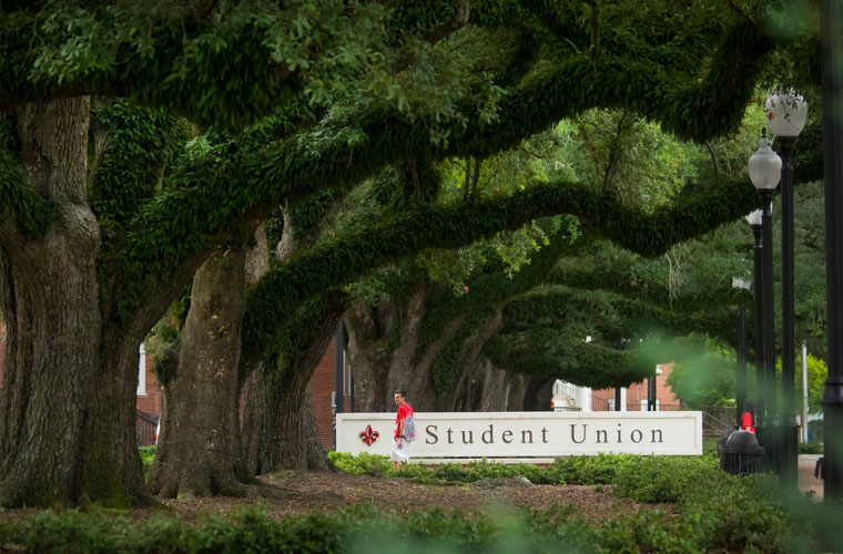 Student walking in front of the 鶹ҹ Student Union sign under a canopy of oak tree branches